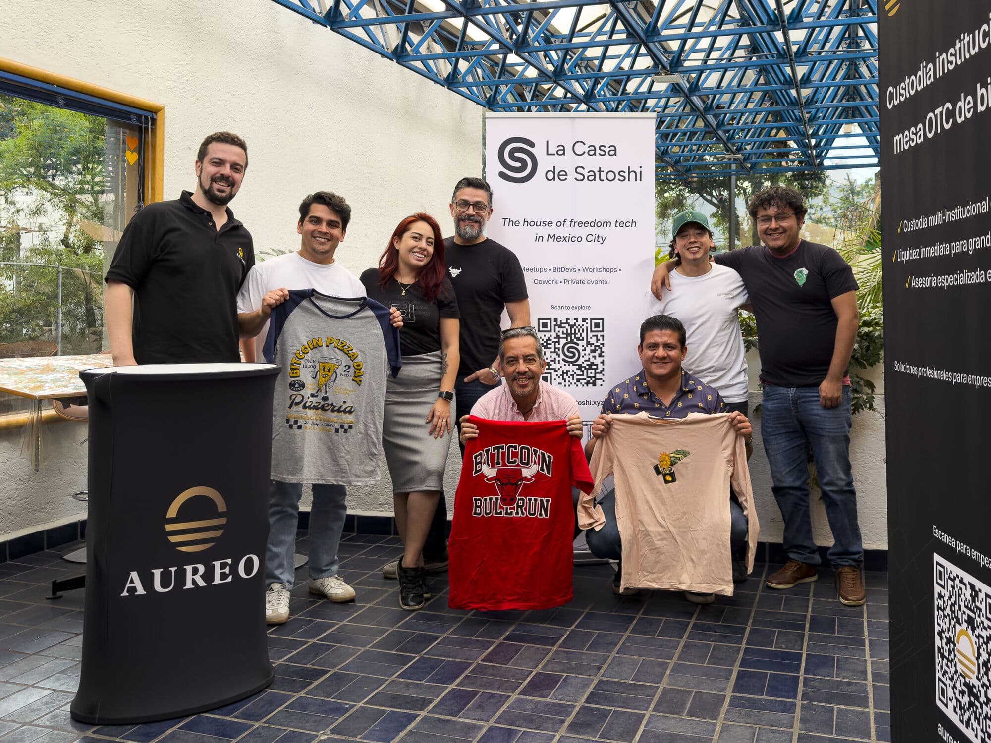 Participantes del hackathon con playeras frente al banner de La Casa de Satoshi